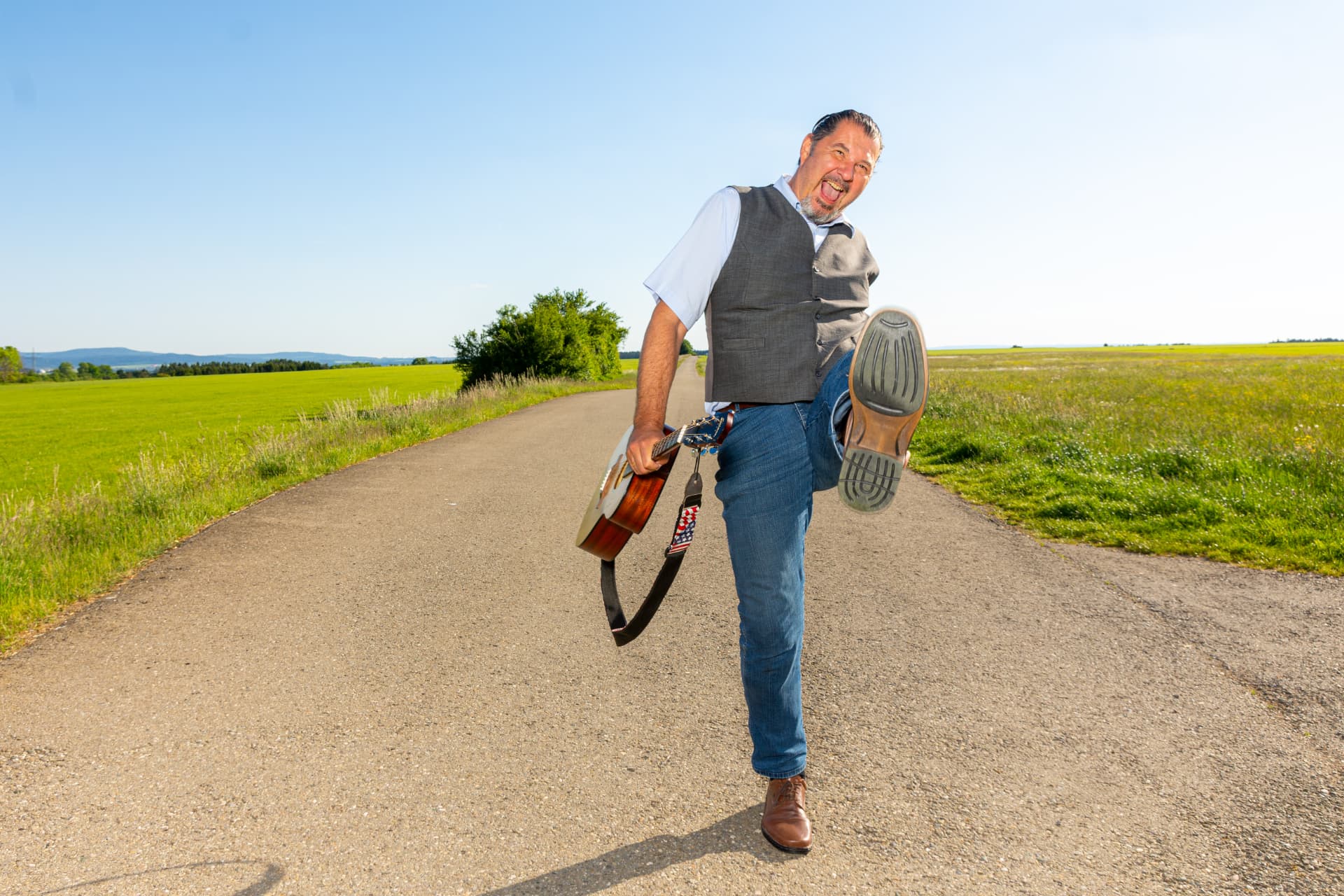 Michael Edel mit Gitarre und schwungvoll angehobenem Bein auf einem Feldweg